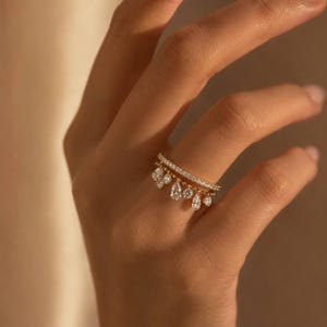 May include: A gold ring with a delicate design, featuring a row of small, sparkling diamonds and a second band with dangling teardrop-shaped gems. The ring is worn on a finger, showcasing its elegant and intricate details.
