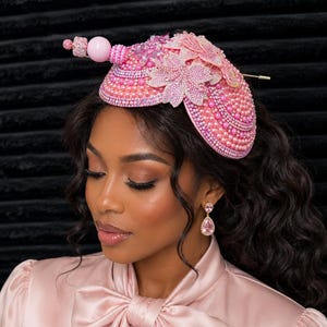 May include: A pink fascinator hat adorned with numerous small pearls, sparkling crystals, and layered floral appliques. A decorative pin featuring a large smooth pink bead and smaller textured beads extends from the side of the structured headpiece.