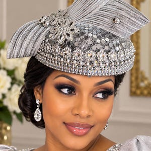 May include: A silver fascinator hat featuring a structured, pleated bow element, a crown covered in reflective circular sequins, and a band of clear, faceted crystal embellishments arranged in a floral pattern around the base of the headpiece.
