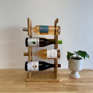 May include: A wooden wine rack holding four bottles of wine. The rack is made of light-colored wood and has a simple, modern design. The bottles are of various colors and labels. A small potted plant sits to the right of the rack.