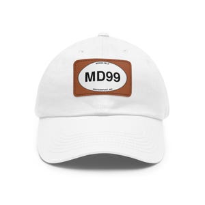 May include: A white baseball cap featuring a rectangular brown leather patch on the front. The patch contains an oval white graphic with the black text MD99, along with the smaller text MORAN FIELD and WESTERNPORT, MD printed on the oval border.