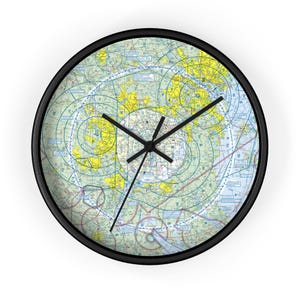 May include: A round wall clock featuring a black frame and a clock face printed with a detailed aviation sectional chart. The map displays complex navigational lines, yellow land masses, and blue water areas, overlaid with black clock hands.
