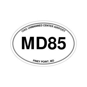 May include: An oval-shaped sticker with a white background and a thin black border. The text OAG UNMANNED CENTER AIRPORT is printed in black along the top curve, MD85 is in large black letters in the center, and PINEY POINT, MD is at the bottom.