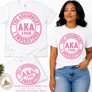 Può includere: Una t-shirt bianca a maniche corte con una grafica circolare rosa che riporta il testo THE ASSIGNMENT I UNDERSTOOD, AKA e 1908. Il design è centrato sul petto. La maglietta è mostrata sia distesa che indossata con jeans blu.