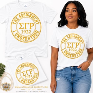 Può includere: Una t-shirt bianca a maniche corte con una grafica circolare dorata che presenta le lettere greche ΣΓΡ, l'anno 1922 e il testo THE ASSIGNMENT I UNDERSTOOD. Il design è centrato sul petto della maglietta.