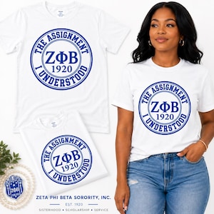 Può includere: Una t-shirt bianca a maniche corte con una grafica circolare blu che riporta il testo "THE ASSIGNMENT I UNDERSTOOD" attorno alle lettere greche "ZΦB" e all'anno "1920". Il design è centrato sul petto del capo.