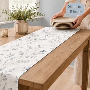 May include: A white table runner featuring a repeating pattern of watercolor-style blue and grey leaves and berries. The runner is laid flat across a wooden table, accompanied by a ribbed ceramic vase with greenery and a stack of beige plates.