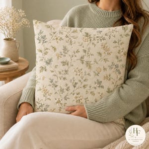 May include: A square throw pillow featuring a cream-colored fabric base with an intricate botanical pattern of small flowers and leaves in muted shades of sage green, dusty blue, and soft yellow, creating a delicate and detailed floral design.