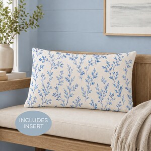 May include: A rectangular throw pillow featuring a cream-colored fabric base with a repeating blue botanical pattern of delicate branches and leaves. The pillow includes an insert and is displayed on a wooden bench with a light blue wall background.