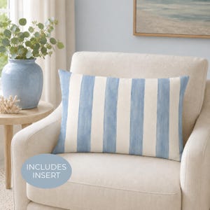 May include: A rectangular throw pillow featuring vertical stripes in light blue and off-white. The fabric has a soft, painted appearance with visible brushstroke textures. A circular light blue badge on the lower left displays the text INCLUDES INSERT.