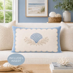 May include: A rectangular lumbar pillow featuring a light blue scalloped border and a central cream-colored panel. The design includes a blue seashell graphic flanked by two beige leafy sprigs. The pillow includes an insert for added structure.