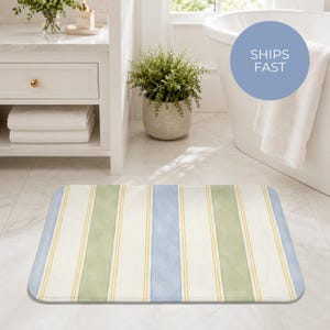 May include: A rectangular bath mat featuring a vertical striped pattern in alternating bands of soft blue, sage green, and cream, accented by thin yellow pinstripes. The mat has rounded corners and a flat, uniform texture across its entire surface.