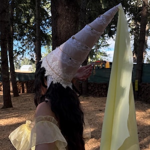 May include: A tall, conical hat made of light-colored fabric with white lace trim and a long, flowing pale yellow veil. The hat features a spiral design with small metallic accents. It is worn with a pale yellow off-the-shoulder top and a pink skirt.