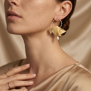 May include: Gold-coloured earrings featuring a distinctive fan-shaped design, each with a dangling pearl. The earrings are attached to a circular hoop. The jewellery is worn on a person's ear. The background is a neutral colour.