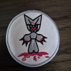 May include: Embroidered patch featuring a gray creature with a menacing expression and red accents. The design includes a white background with a gray border and a splash of red at the bottom, creating a bold and edgy aesthetic.