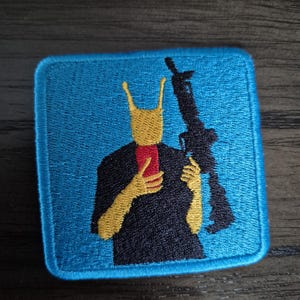 Puede incluir: Un parche bordado cuadrado con un fondo azul brillante. La figura central tiene una cabeza amarilla con antenas, viste una camiseta negra y sostiene un teléfono inteligente rojo en una mano y un rifle negro en la otra.