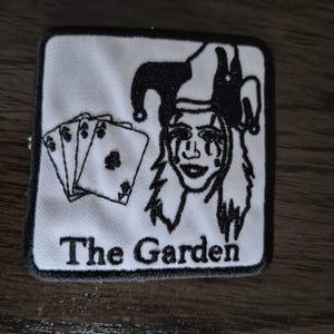 以下が含まれることがあります： 白い背景に黒い縁取りが施された正方形の刺繍ワッペン。メイクをした道化師の顔の黒い線画イラスト、4枚のトランプのカード、そして下部に黒字で「The Garden」というテキストがデザインされています。