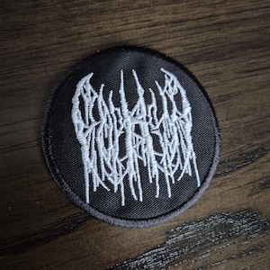 May include: A circular black embroidered patch featuring white thread stitched in a dense, jagged, and vertical style that resembles illegible, stylised lettering or a complex abstract pattern centred on the fabric surface.