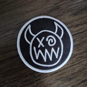 May include: A round, black patch with a white embroidered design. The design features a cartoonish face with horns, an 'X' and 'o' for eyes, and a zig-zag mouth. The patch has a white border.