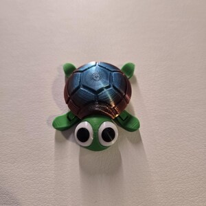 May include: A 3D-printed turtle figurine with a green body, four flippers, and a segmented shell in shades of blue and brown. The turtle has large, googly eyes. This decorative item is likely a toy or collectible.