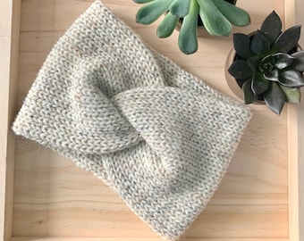 May include: A light gray knitted headband with a twisted knot design. The headband is on a wooden surface with two potted succulents.