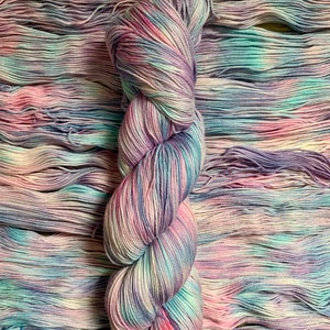 May include: A skein of hand-dyed yarn in a blend of pastel colors, including pink, purple, blue, and white. The yarn is arranged on a wooden surface, with the skein in the center and loose strands surrounding it. The yarn appears soft and textured.