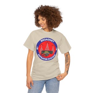 Puede incluir: Una camiseta color beige con un diseño circular que dice "BROADNET REPEATER SYSTEMS". El diseño presenta un fondo rojo, un borde azul e ilustraciones de torres de radio y una radio.