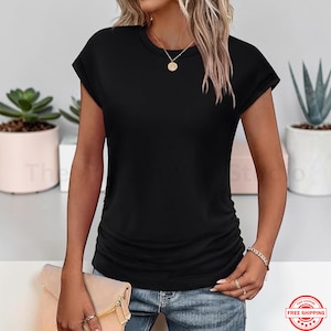 May include: A black short-sleeved t-shirt with a round neckline. The shirt has a fitted silhouette with ruching on the side. The model is wearing blue jeans and a gold necklace. A clutch is held in the hand.