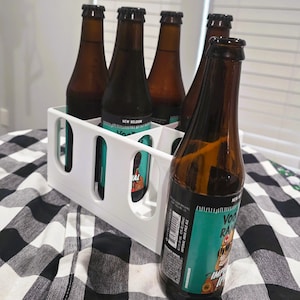 May include: A white plastic beer bottle carrier holding five brown glass beer bottles. The bottles have black caps and teal and black labels. The carrier sits on a black and white checkered surface.