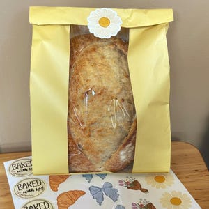 May include: A loaf of golden-brown sourdough bread packaged in a light yellow paper bag with a clear plastic window. The bag is sealed at the top with a white daisy sticker. Below the bag is a sheet of stickers featuring croissants and flowers.