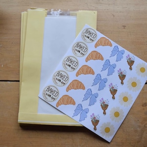 May include: A stack of yellow paper bags with a clear window, next to a sheet of stickers. The stickers feature the text "BAKED with love" in a circle, along with croissant, bow, flower bouquet, and daisy designs.