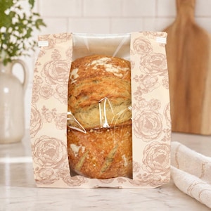 May include: Two loaves of freshly baked bread are visible through a clear window in a floral-patterned paper bag. The bag has a beige background with a repeating pattern of pink flowers. The bread has a golden-brown crust.