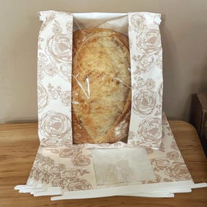 Può includere: Una pagnotta di pane a lievitazione naturale dorato all'interno di un sacchetto di carta rettangolare con un motivo floreale beige chiaro. Il sacchetto ha una finestra in plastica trasparente sul davanti, che rende visibile il pane croccante.