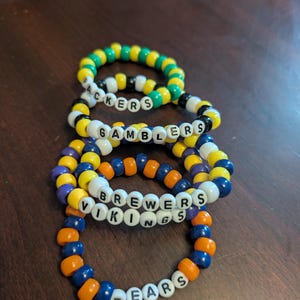 May include: Four beaded bracelets with team names: Packers, Gamblers, Brewers, Vikings, and Bears. The bracelets feature a variety of colors including green, yellow, purple, orange, and blue. The letters are white.