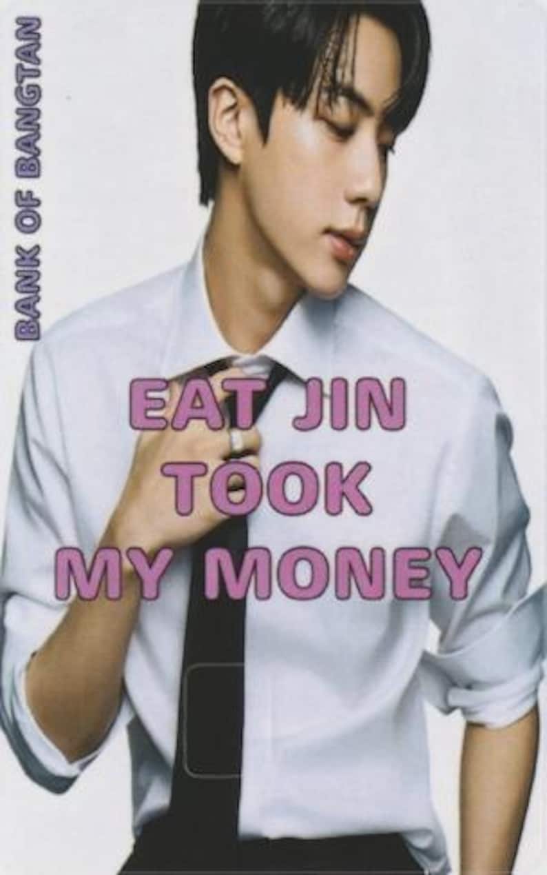 以下が含まれることがあります： ピンク色の「EAT JIN TOOK MY MONEY」の文字が入ったカード。白いシャツと黒いネクタイをした人物が写っています。「BANK OF BANGTAN」の文字が左側に縦書きで印刷されています。
