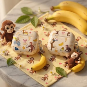 May include: Two white AirPod cases with a repeating pattern of bananas, monkeys, and stars. One case has a monkey riding a bicycle. A plush monkey sits next to each case. A yellow banana and a yellow patterned cloth are also in the image.