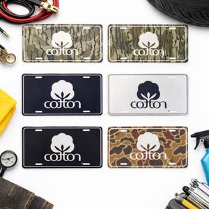 May include: Six license plates featuring a cotton logo. Two plates have a camouflage pattern, one is black, one is silver, and one is black with a white border. The logo is a white cotton boll with the word "cotton" below it.