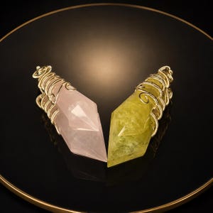 May include: Two faceted crystal pendants, one pale pink and one translucent yellow, each secured with gold-toned wire wrapping at the top. The pendants are pointed at the base and feature a coiled wire loop for suspension at the top of each stone.