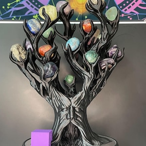 May include: A black tree-shaped sculpture with a face carved into the trunk, featuring multiple branches holding various polished gemstones in colors including blue, green, orange, purple, and metallic gold, next to a small purple cube.