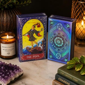 May include: Two rectangular tarot card decks in plastic wrap. The left deck features The Fool card illustration with a yellow background, a figure, and a dog. The right deck has a blue and teal holographic mandala design. Both are upright.