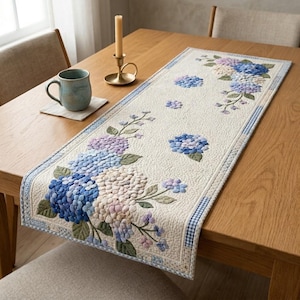 May include: Cream-colored table runner with embroidered blue, purple, and white hydrangea flowers. The runner has a blue and white gingham border and is placed on a wooden table. A mug, coaster, candle, and candlestick are also on the table.