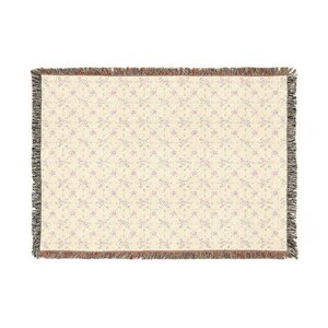 May include: Cream-colored woven throw blanket with a repeating floral pattern in light purple. The blanket has a fringed edge in shades of brown, black, and cream. The floral design adds a touch of elegance to this home decor item.