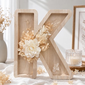 Könnte beinhalten: Ein Holzbuchstabe "K", verziert mit cremefarbenen Blumen und Blättern. Der hellbraune Buchstabe hat eine vertiefte Mitte, wodurch ein Schatteneffekt entsteht. Die Blumengestecke beinhalten eine große, mehrschichtige Blume und kleinere Blüten.