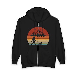 May include: A black zip-up hoodie featuring a graphic print of a silhouette of Bigfoot walking a dog on a leash. The design includes a retro-style sunset background with horizontal stripes in orange, yellow, and teal, topped by a mountain range.