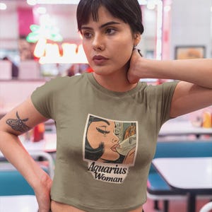 May include: Olive green crop top featuring a comic-style graphic of a woman with the text "Aquarius Woman". The graphic is in a square frame. The top is short-sleeved and worn by a person.