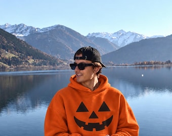 May include: A bright orange hooded sweatshirt featuring a black jack-o'-lantern face graphic on the front. The garment has long sleeves, a hood, and a front kangaroo pocket. The wearer is also sporting a black baseball cap and dark sunglasses.