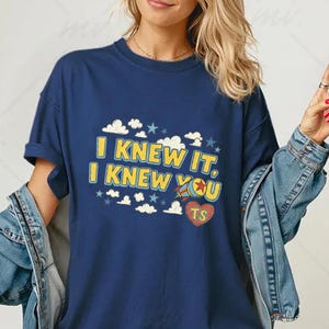May include: A navy blue short-sleeve t-shirt featuring the yellow text "I KNEW IT, I KNEW YOU" with a small rocket ship graphic, a red heart containing the letters "TS", and scattered white clouds and blue stars printed on the front chest area.