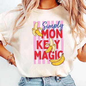 May include: A cream-colored short-sleeve t-shirt featuring a graphic print with vertical pink stripes. The text reads Simply MON KEY MAGIC in blue and red letters, accompanied by three yellow banana illustrations arranged around the words.