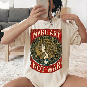 以下が含まれることがあります： 詳細なグラフィックが施されたクリーム色のTシャツ。デザインには、赤い文字で「MAKE ART NOT WAR」、女性の横顔、鳩が含まれています。アートワークは、金と緑のアクセントで円形のデザインにフレームされています。