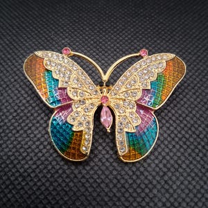 May include: A gold-toned butterfly brooch with a colorful, rainbow-hued enamel design on the wings. The brooch is embellished with clear crystals and pink gemstones. The butterfly's body is black.